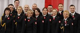 Grupa funkcjonariuszy PSP w galowych mundurach stoi razem do zdjęcia zbiorowego na tle biało-czerwonej dekoracji; wszyscy mają przypięte biało-czerwone rozety