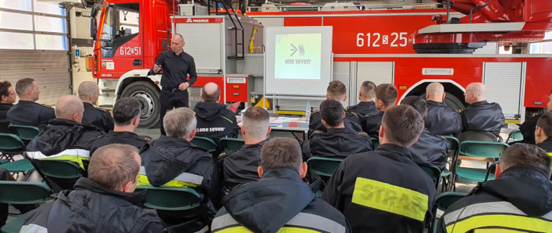 Na pierwszym planie odwróceni plecami strażacy, ubrani w czarne kurtki z napisem Straż. Na drugim planie mężczyzna prowadzący szkolenie ubrany w czarną koszulę i czarne spornie, w tle samochód ratowniczo - gaśniczy 