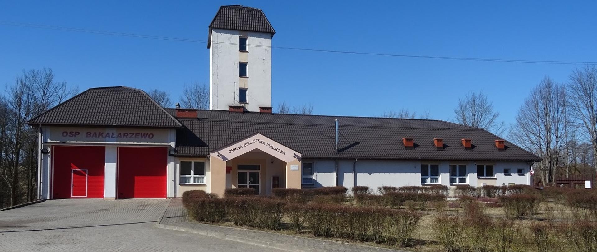 Widoczny jest budynek o ciemny dachu, jasnymi ścianami, czerwonymi dwiema bramami i wieżą. Przed budynkiem podjazd z kostki brukowej a po prawej stronie żywopłot.