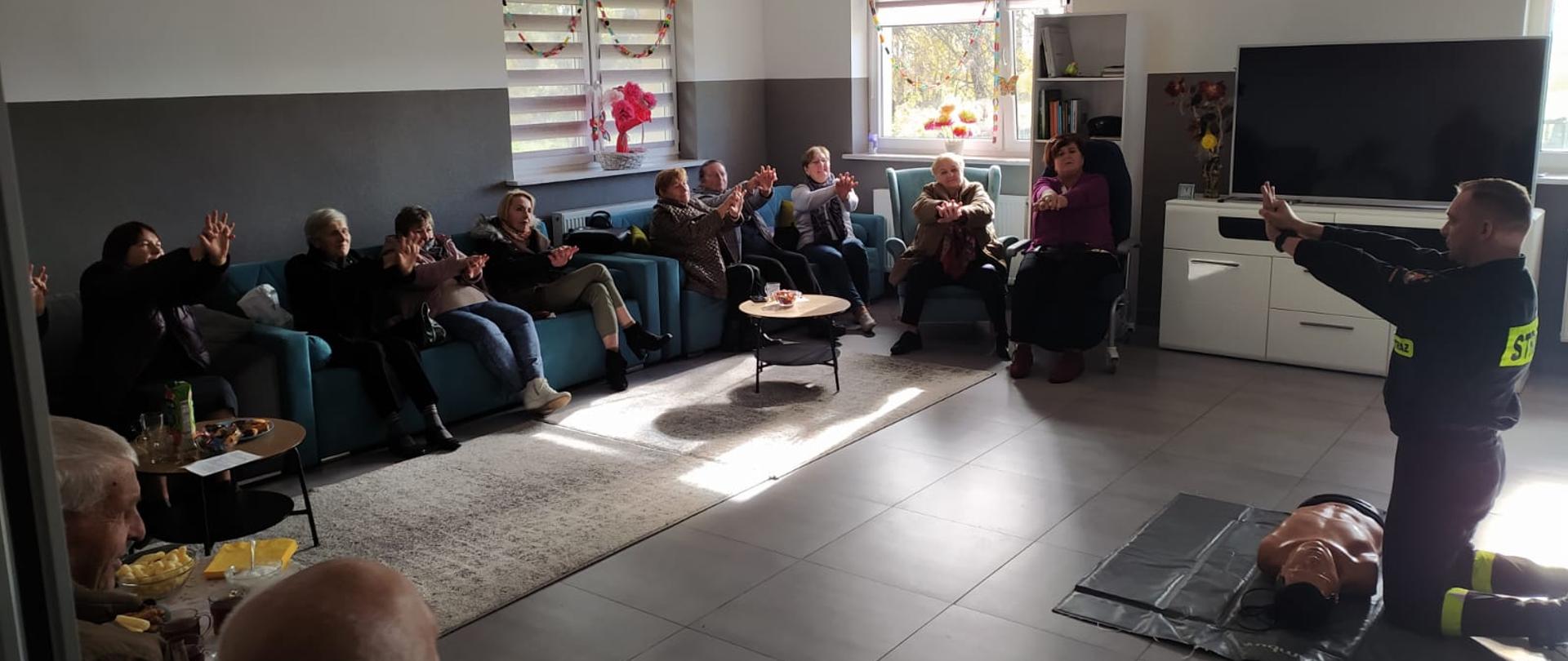 W pomieszczeniu przed fantomem klęczy strażak w czarnym mundurze. Trzyma ręce złączone przed sobą. Prze nim siedzi kilkanaście seniorek trzymających ręce w ten sam sposób.