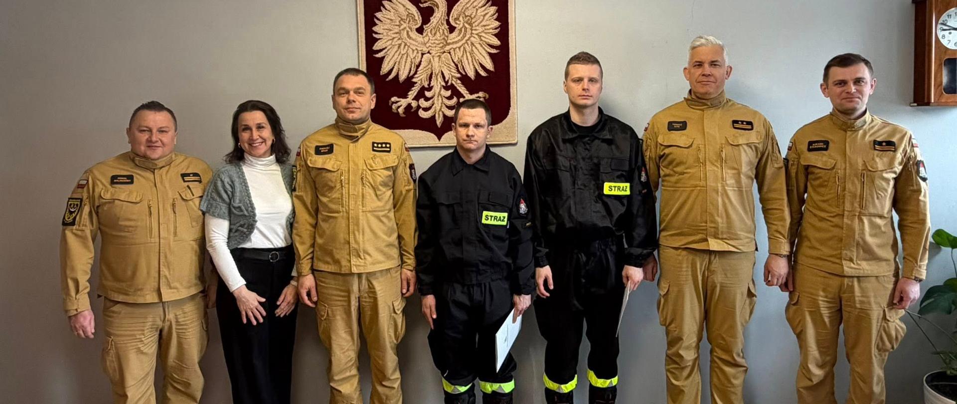 W pomieszczeniu urzędowym siedem osób stoi w szeregu — pięć w beżowych mundurach służbowych oraz dwie w czarnych uniformach strażackich z odblaskowym napisem. Za nimi widnieje duży herb z białym orłem, a obok znajduje się zegar i roślina w donicy. Osoby pozują do wspólnego zdjęcia z okazji ślubowania nowych strażaków.