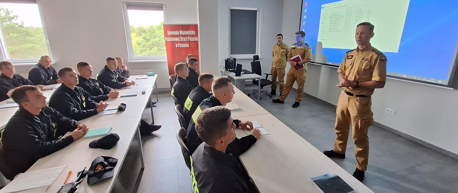 sala szkoleniowa na której siedzą strażacy, przed nimi stoi trzech strażaków, duży ekran 
