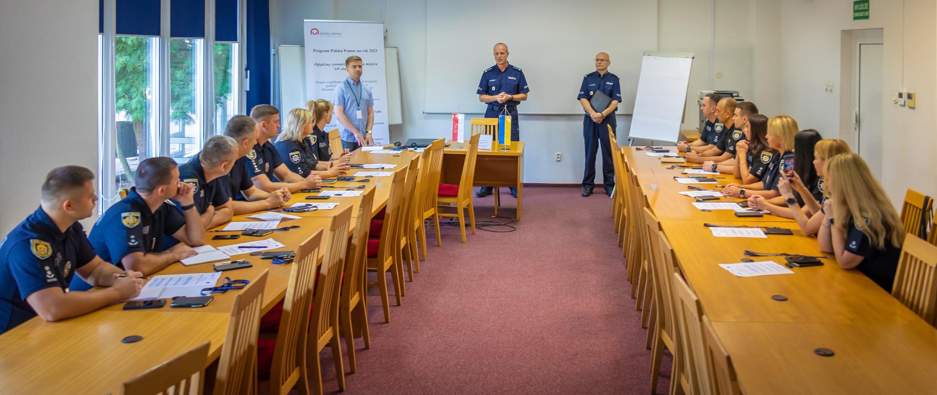 W sali lekcyjnej grupa mężczyzn i kobiet siedzi przy ustawionych naprzeciwko siebie dwóch rzędach stołów i patrzy się na prowadzących szkolenie trzech mężczyzn stojących przed pustym ekranem, tablicą flipchart i banerem z logo Polskiej pomocy