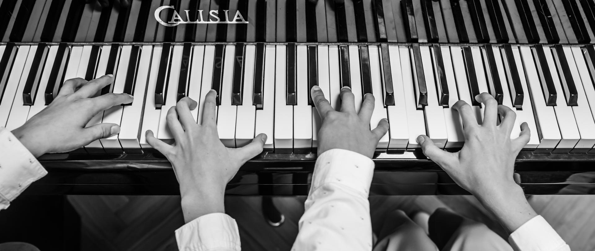 czarno-białe zdjęcie rąk dwóch osób grających na fortepianie, perspektywa z lotu ptaka, dłonie odbijają się w desce fortepianu