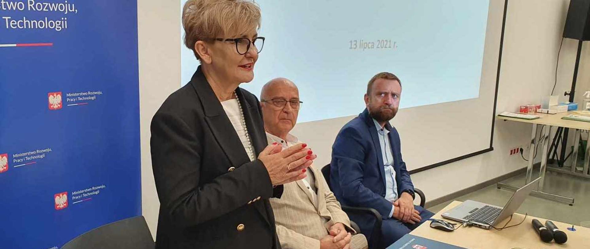 Wiceminister Iwona Michałek stoi przed stołem i uczestniczy w konsultacjach dotyczących sytuacji na rynku pracy w Polsce. Obok niej na kszesłach siedzi dwóch ekspertów. 