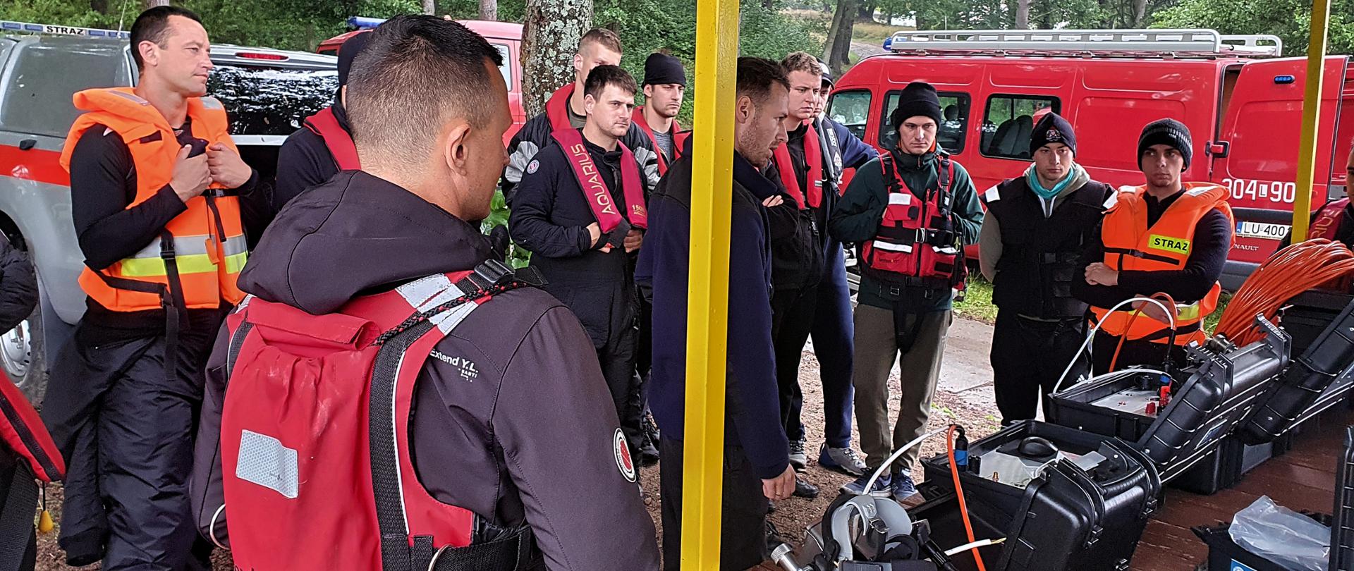 Grupa strażaków uczestniczących w szkoleniu stojąca pod metalową wiatą, gdzie na stoliku prezentowany jest sprzęt łączności wykorzystywany podczas prac podwodnych. W tle widoczne są dwa strażackie samochody. 