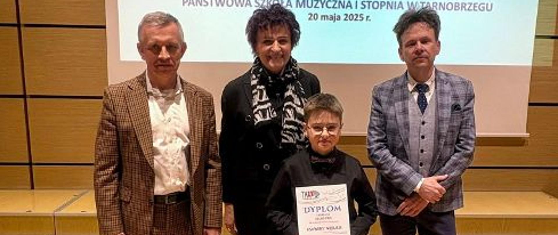Zdjęcie przedstawia ucznia trzymającego dyplom laureata oraz Jury konkursu. Z tyłu widoczny plakat promujący Tarnobrzeski Konkurs Akordeonowy.