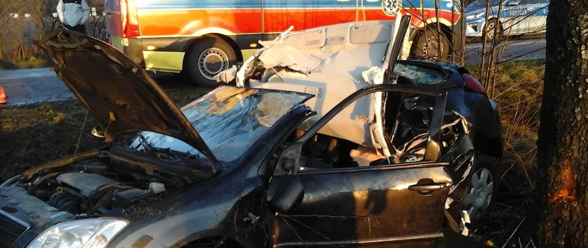 Samochód osobowy po uderzeniu w drzewo stoi na poboczu jezdni. W aucie odcięto dach. W tle karetka pogotowia ratunkowego i radiowóz.