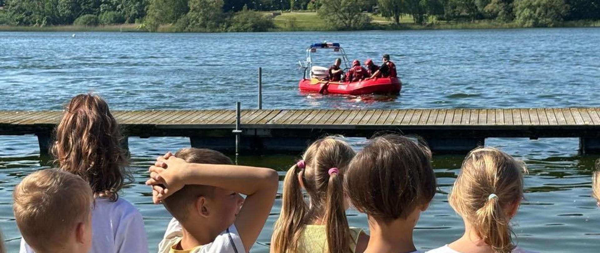 dzieci stojące tyłem do kadru a przed nimi łódka z ratownikami na jeziorze