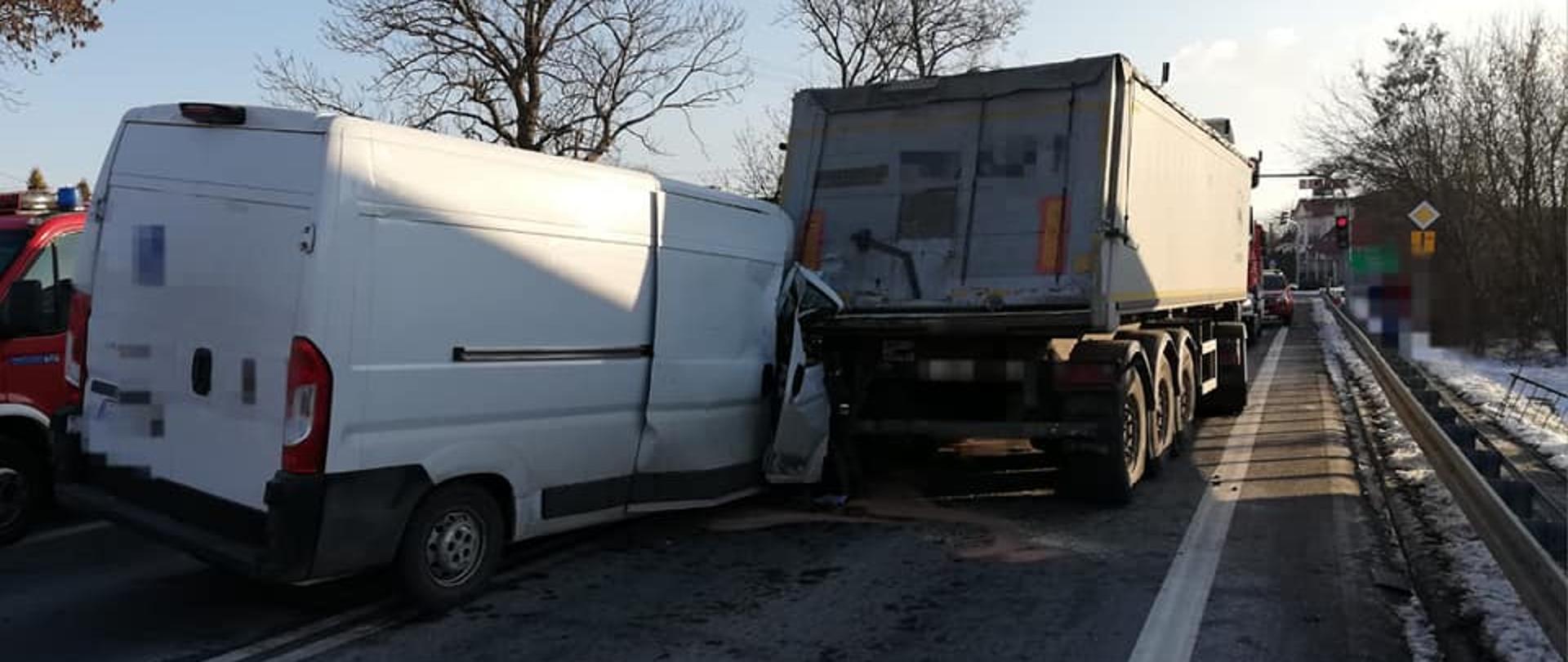 Na zdjęciu widać białego busa, który uderzył w tył naczepy samochodu ciężarowego. Oba samochody stoją na środku asfaltowej jezdni przed skrzyżowaniem z sygnalizacją świetlną. Zza samochodów widać czerwone samochody strażackie. W tle widać budynki i drzewa. Na poboczu leży śnieg. Jest widno. 