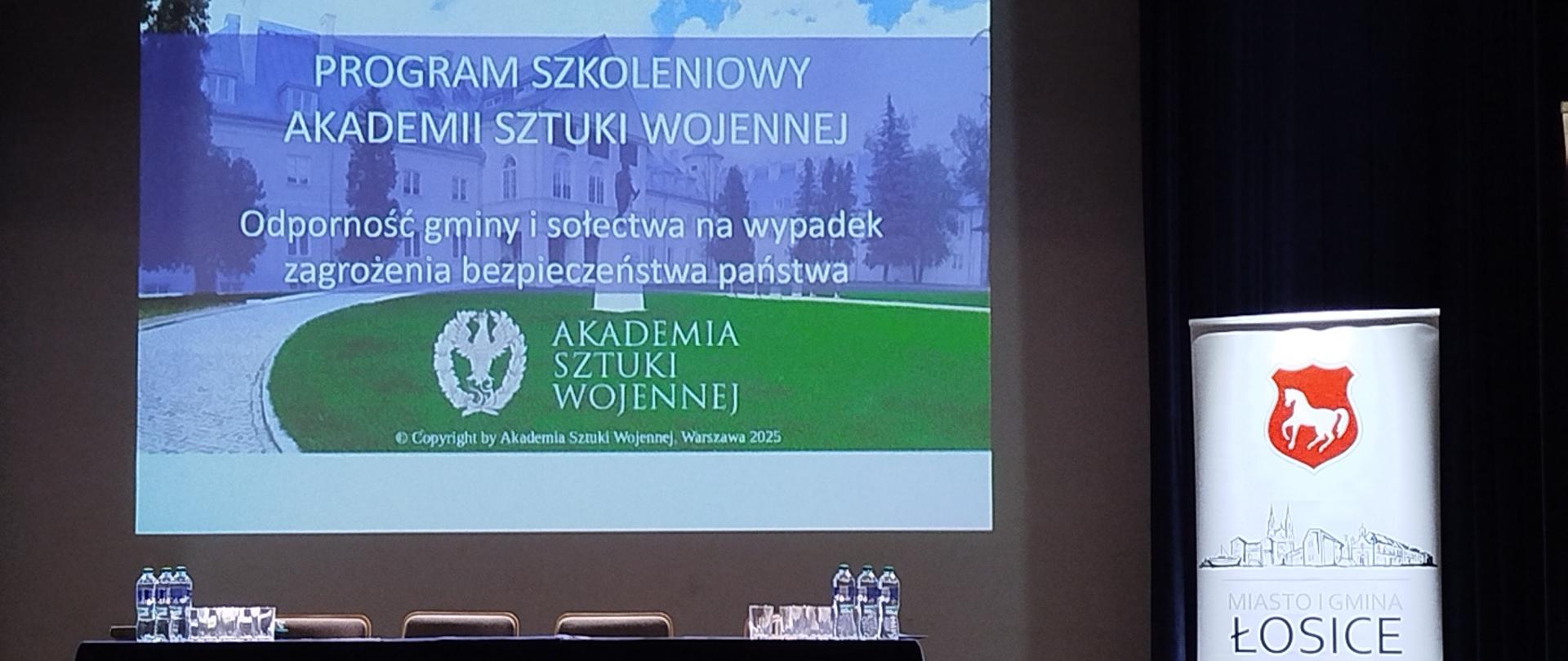 Na zdjęciu widać scenę z ustawionym stołem i czterema krzesłami, przeznaczonymi dla prelegentów szkolenia, za stołem na ekranie wyświetlona prezentacja, po prawej stoi rollup z herbem miasta gminy Łosice