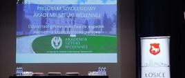 Na zdjęciu widać scenę z ustawionym stołem i czterema krzesłami, przeznaczonymi dla prelegentów szkolenia, za stołem na ekranie wyświetlona prezentacja, po prawej stoi rollup z herbem miasta gminy Łosice
