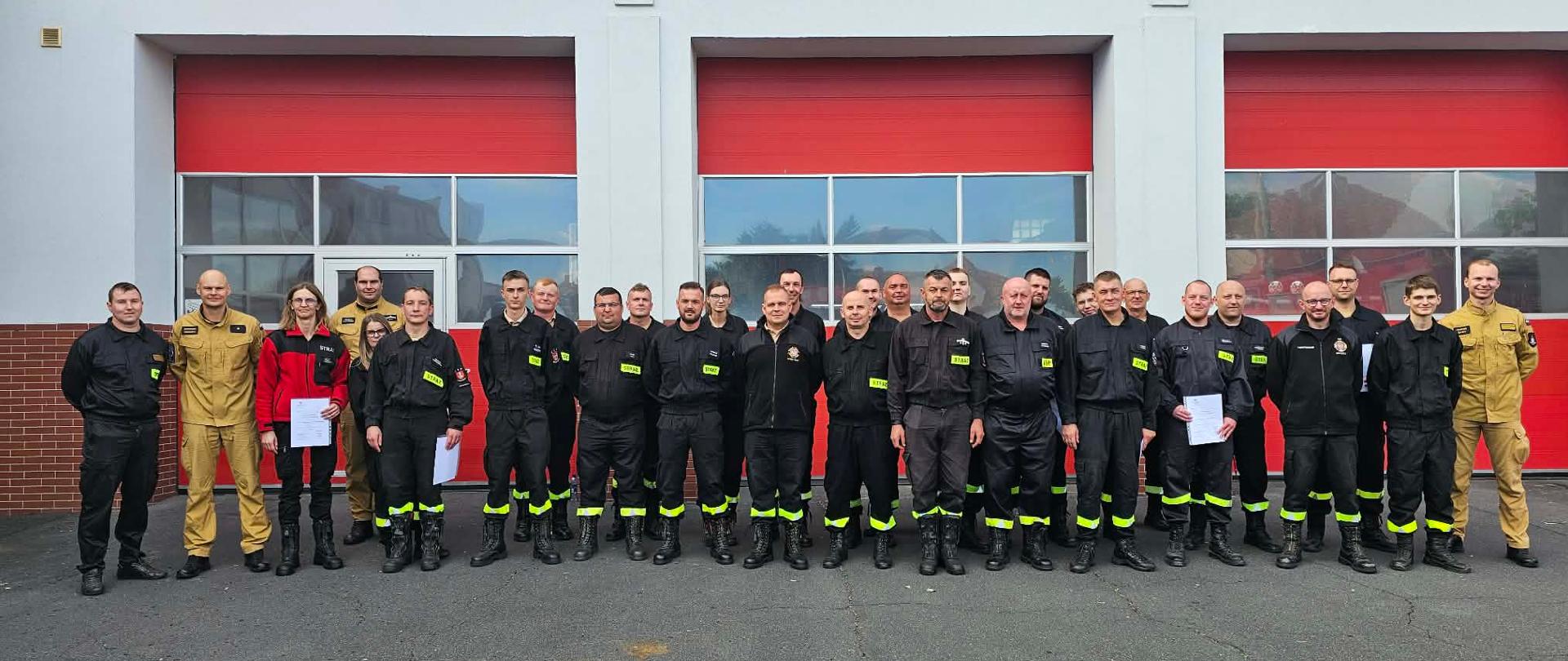 Druhów oraz strażaków z komisji egzaminacyjnej. Stoją oni przed budynkiem komendy, na placu wewnętrznym i pozują do zdjęcia. W rękach trzymają zaświadczenia potwierdzające ukończenie z wynikiem pozytywnym egzaminu.