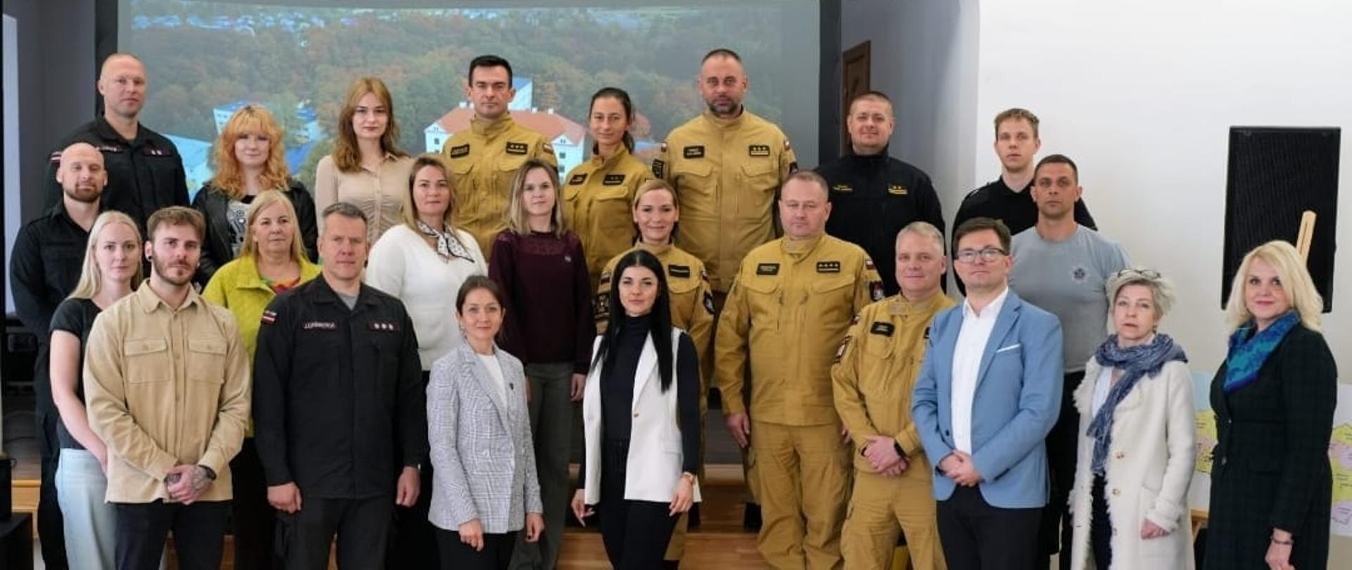 Wspólna fotografia uczestników polsko-łotewskiej wymiany w sali z ekranem; w pierwszym rzędzie stoją przedstawiciele PSP i SFRS oraz organizatorzy.