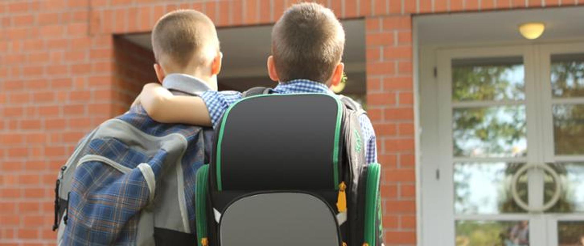 Po prawej stronie chłopiec w koszuli, który niesie czarno-zielony plecak i obejmuje drugiego chłopca niosącego na plecach szaro-granatowy plecak