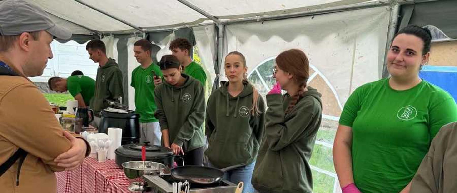 Zdjęcie przedstawia plenerowe stoisko gastronomiczne pod białym namiotem ogrodowym, przy którym grupa młodych ludzi przygotowuje jedzenie.Po prawej stronie, za podłużnym stołem nakrytym obrusem w biało-czerwoną kratkę, stoi siedmioro nastolatków lub młodych dorosłych. Większość z nich jest ubrana w pasujące do siebie, ciemnozielone bluzy z kapturem (hudie) z małym, okrągłym logo na piersi. Jedna z dziewczyn po prawej stronie ma na sobie jasnozielony t-shirt z tym samym logo oraz różowe, ochronne rękawiczki nitrylowe. Młodzi ludzie rozmawiają ze sobą i zajmują się obsługą stanowiska.Na stole rozstawiony jest sprzęt gastronomiczny oraz akcesoria kuchenne. Widoczne są m.in. podgrzewacze gastronomiczne, patelnia, gofrownica, papierowe talerzyki, kubki oraz plastikowe sztućce.Po lewej stronie, tyłem do obiektywu, stoi mężczyzna w brązowej kurtce i czapce z daszkiem, który przygląda się pracy na stoisku lub czeka na zamówienie. Całe wydarzenie odbywa się na trawiastym podłożu, a w tle widać fragmenty zieleni.