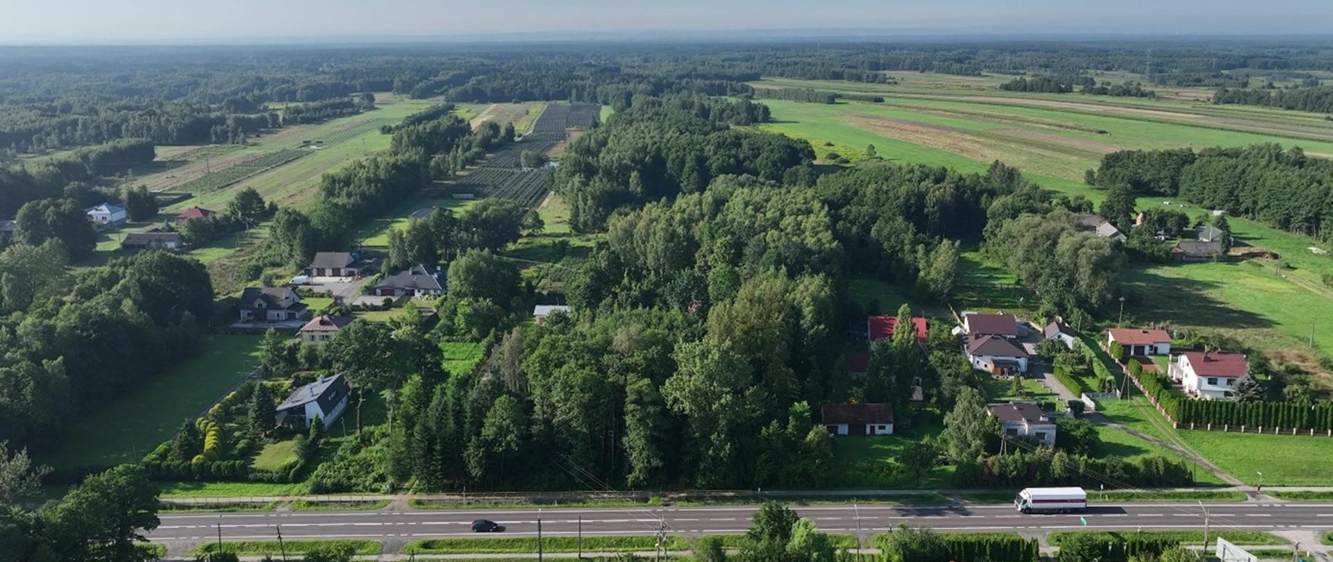 Widok z lotu ptaka na spokojną, wiejską okolicę. Wśród licznych drzew i zielonych pól rozrzucone są pojedyncze domy z ogrodami. Na pierwszym planie biegnie szeroka droga, po której jedzie ciężarówka i samochód osobowy. W tle rozciąga się rozległy, płaski krajobraz pól oraz lasów pod jasnym, bezchmurnym niebem.