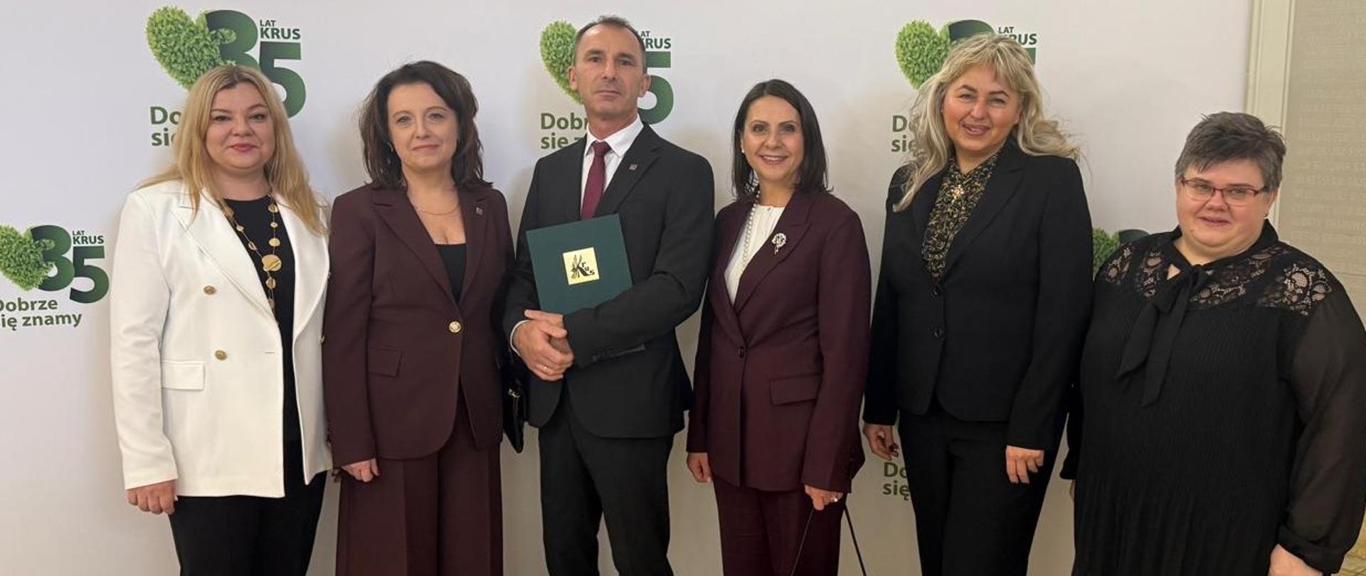 Pięć kobiet i mężczyzna stojący pośrodku grupy pozują do zdjęcia na tle okolicznościowej ścianki na 35-lecie KRUS. Mężczyzna trzyma w rękach zieloną teczkę ze złotym logo KRUS. Wszyscy uśmiechają się do zdjęcia.