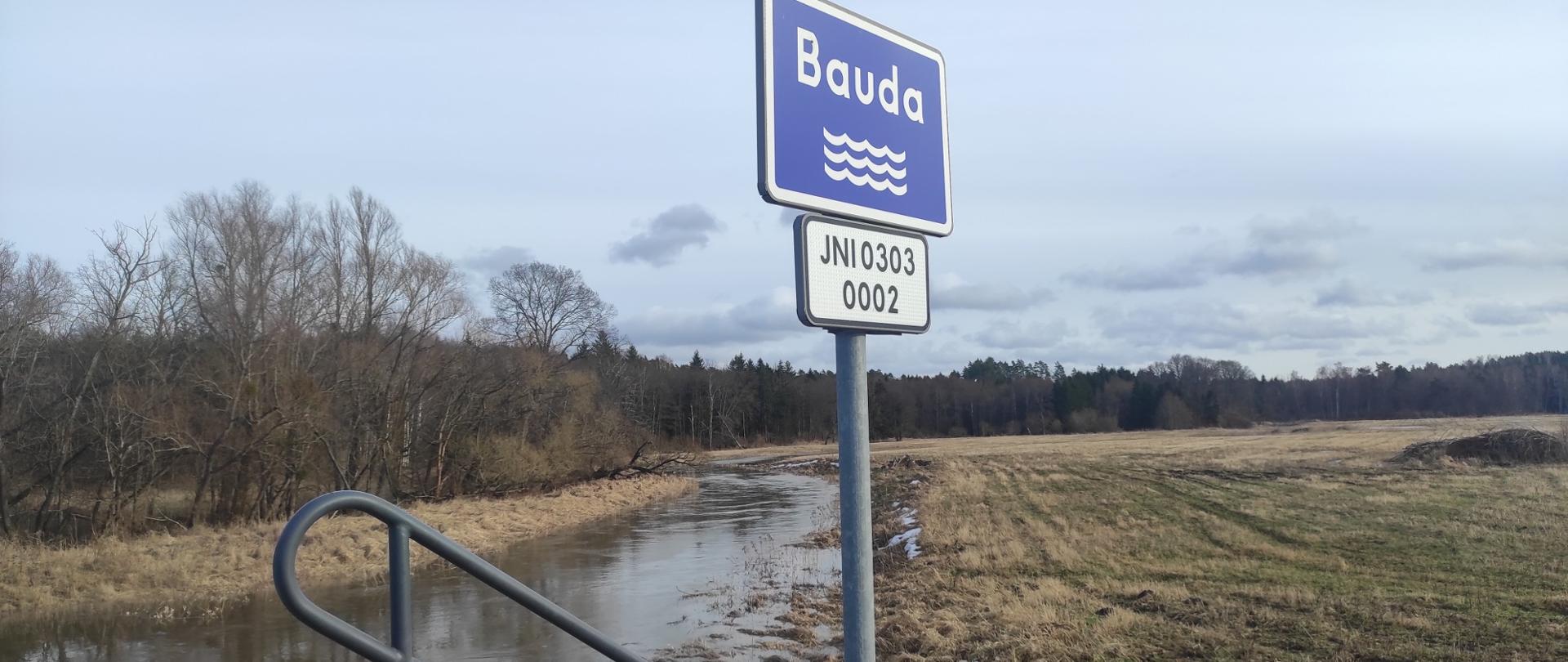 Zdjęcie zrobione nad brzegiem rzeki, wysoki stan wody. Na środku znak z nazwą rzeki Bauda. Pochmurne niebo.