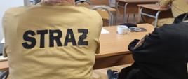 Strażacy siedzą w ławkach podczas szkolenia na ekranie wyświetlana jest prezentacja.