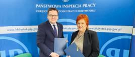 2 osoby na tle banneru w kolorze niebieskim z logo i napisem Państwowa Inspekcja Pracy. Osoby trzymają teczki w kolorze niebieskim. Od lewej strony: Pan dr Karol Łapiński p.o. Okręgowego Inspektora Pracy w Białymstoku, Pani dr n. med. Maria Siewko – Podlaski Państwowy Wojewódzki Inspektor Sanitarny w Białymstoku.
