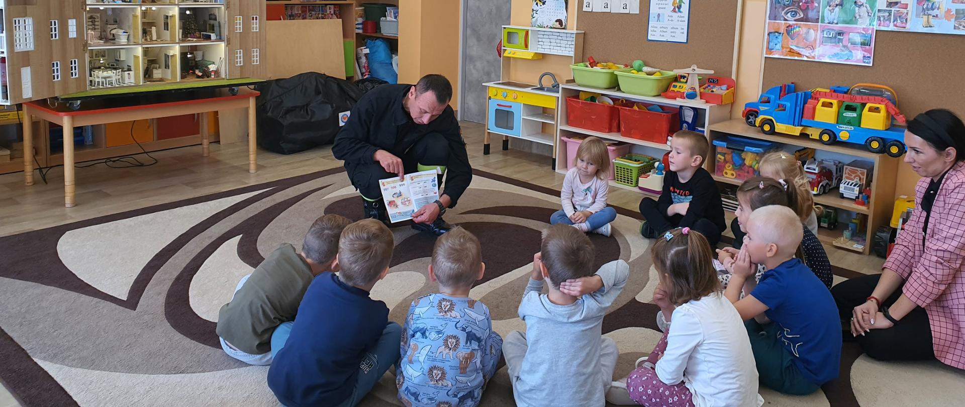 Strażak za pomocą materiałów edukacyjnych omawia numery alarmowe służb ratunkowych. Dzieci przyglądają się książce odpowiadając na zadawane przez strażaka pytania. Działania prewencyjne odbywają się w klasie przedszkolnej. 