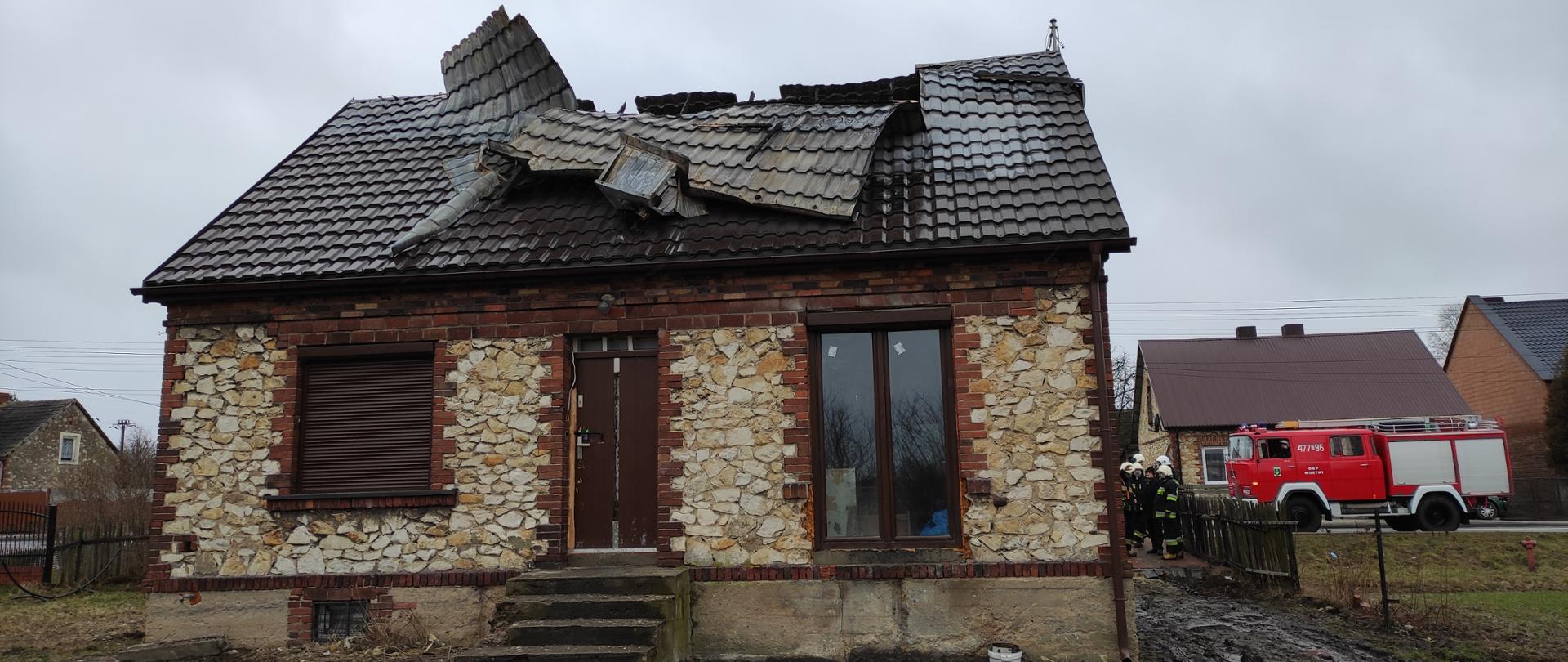 Na zdjęciu widać budynek mieszkalny, murowany z kamienia wapiennego oraz cegły. Dach dwuspadowy z blachy koloru brązowego, Część blachy na dachu została zerwana przez wiatr. W tle widać budynki oraz samochód pożarniczy. 