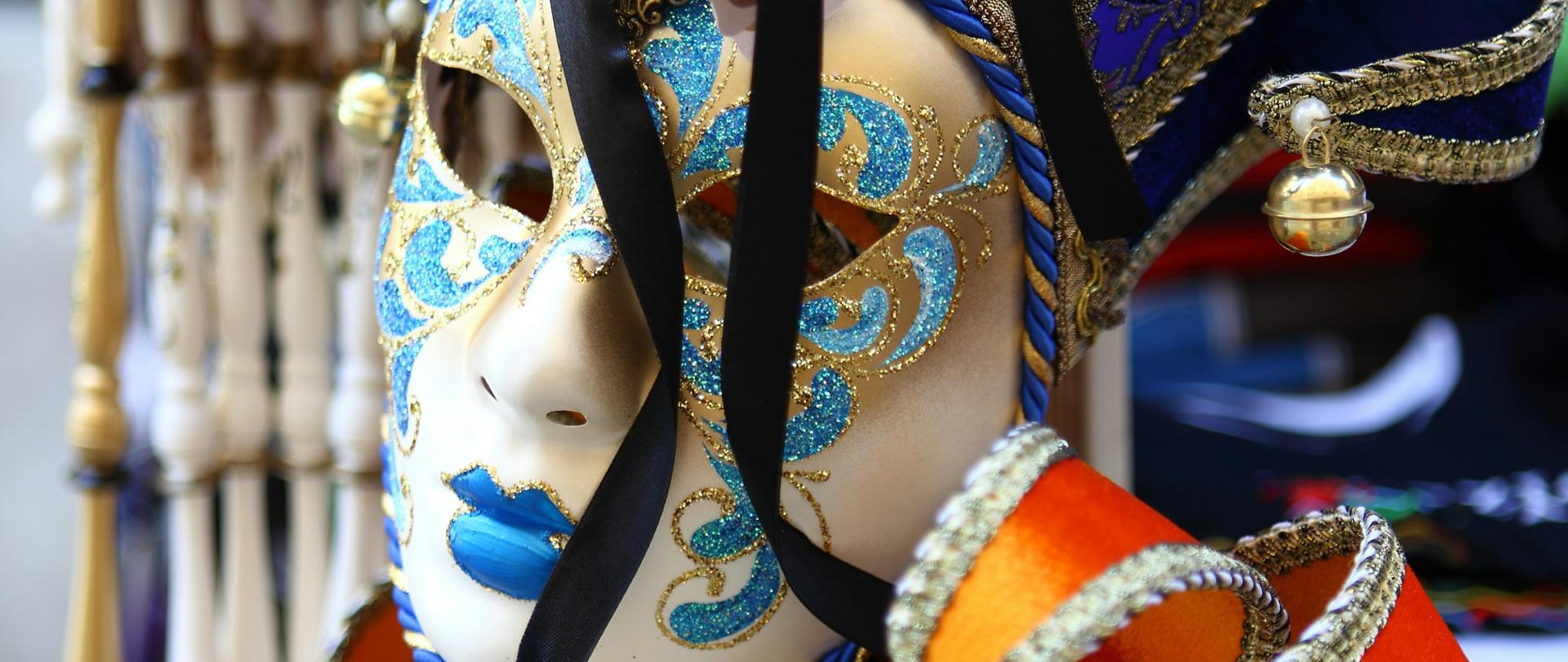 Zbliżenie na weneckie maski karnawałowe bogato zdobione złotymi ornamentami, niebieskimi i pomarańczowymi detalami oraz małymi dzwoneczkami, zawieszone na ekspozycji.