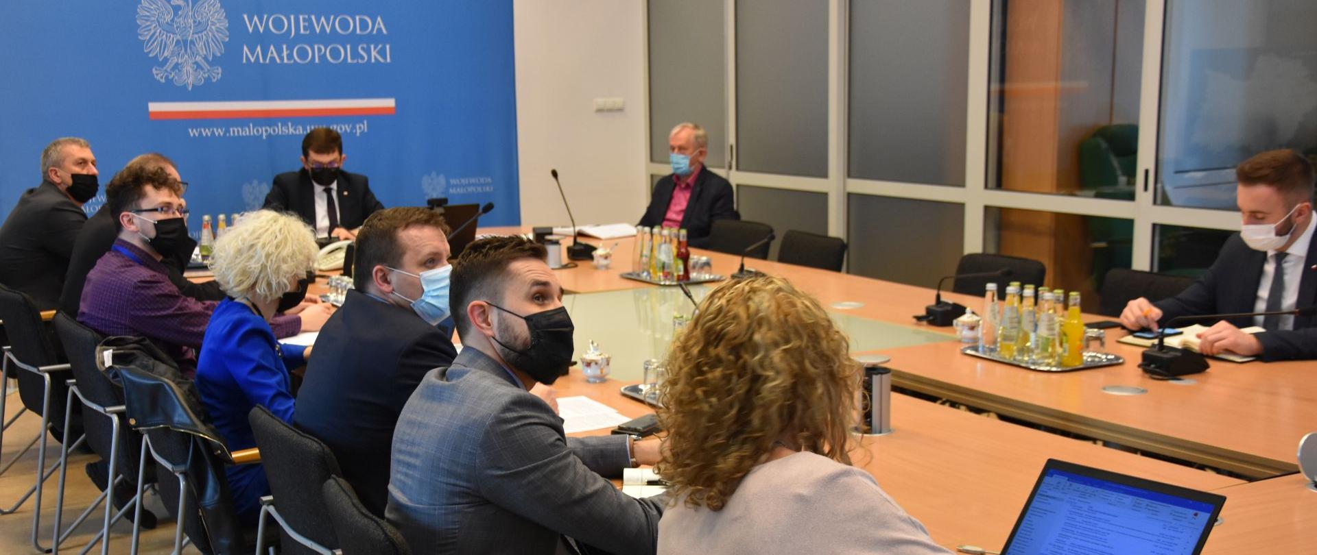 Przy stole siedzi grupa ludzi uczestnicząca w posiedzeniu. Na ścianie niebieski baner z białymi literami WOJEWODA Małopolski.