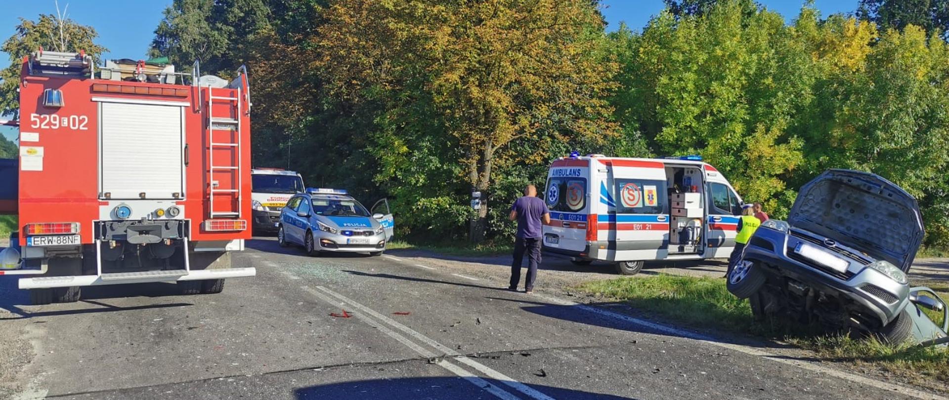 Na lewym pasie drogi asfaltowej stoi samochód strażacki (tyłem do fotografa). Na prawym pasie drogi asfaltowej stoi osobowy samochód policyjny. Na piaszczystej drodze odchodzącej od asfaltowej drogi, tuz przed czołem samochodu policyjnego, stoi karetka, a przed nią w rowie znajduje się srebrny samochód osobowy. Na drodze stoi mężczyzna w czarnych spodniach i ciemnym podkoszulku.