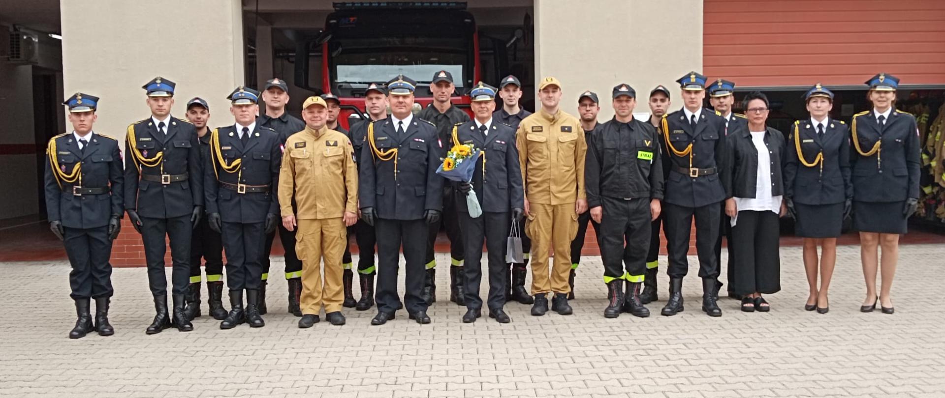 Strażacy wraz z pracownikami cywilnymi przed budynkiem Komendy PSP w Rawiczu. W centralnej części Komendant Powiatowy PSP, po prawej stronie trzymający bukiet nowo mianowany Dowódca Jednostki Ratowniczo-Gaśniczej. W tle bramy wyjazdowe z garażu Komendy oraz napis na elewacji budynku.