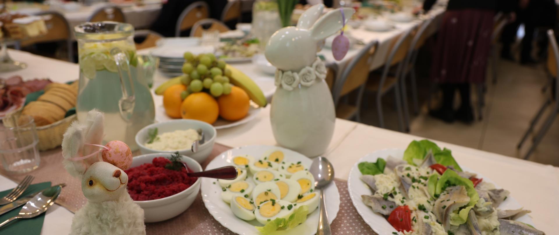 Zbliżenie na fragment stołu przykrytego jasnym obrusem. Na pierwszym planie widać dekoracyjną figurkę puszystego zajączka z różową pisanką. Obok niego półmiski z tradycyjnymi daniami: faszerowane jajka posypane szczypiorkiem, miseczka z ćwikłą oraz śledzie w śmietanie. W tle widać dzbanki z wodą, owoce oraz rozmyte sylwetki gości siedzących przy dalszych stołach w jasnej, przestronnej sali.