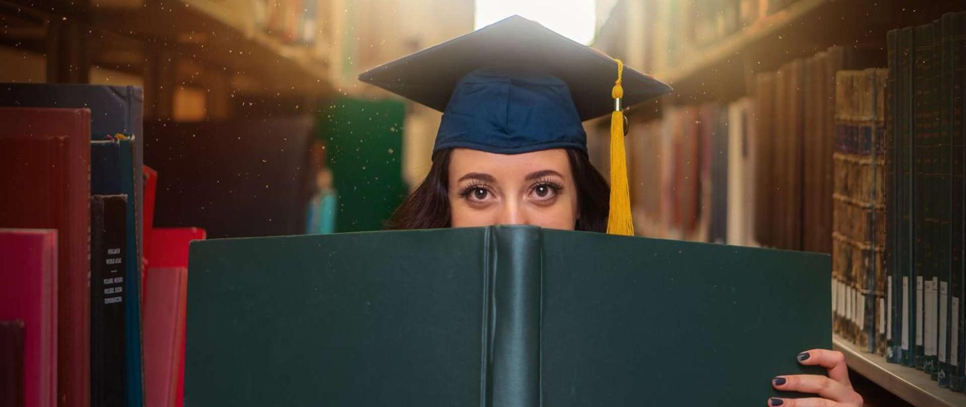 kobieta w granatowym birecie stoi na tle regałów z książkami i trzyma w dłoniach dużą księgę, która zasłania połowę jej twarzy