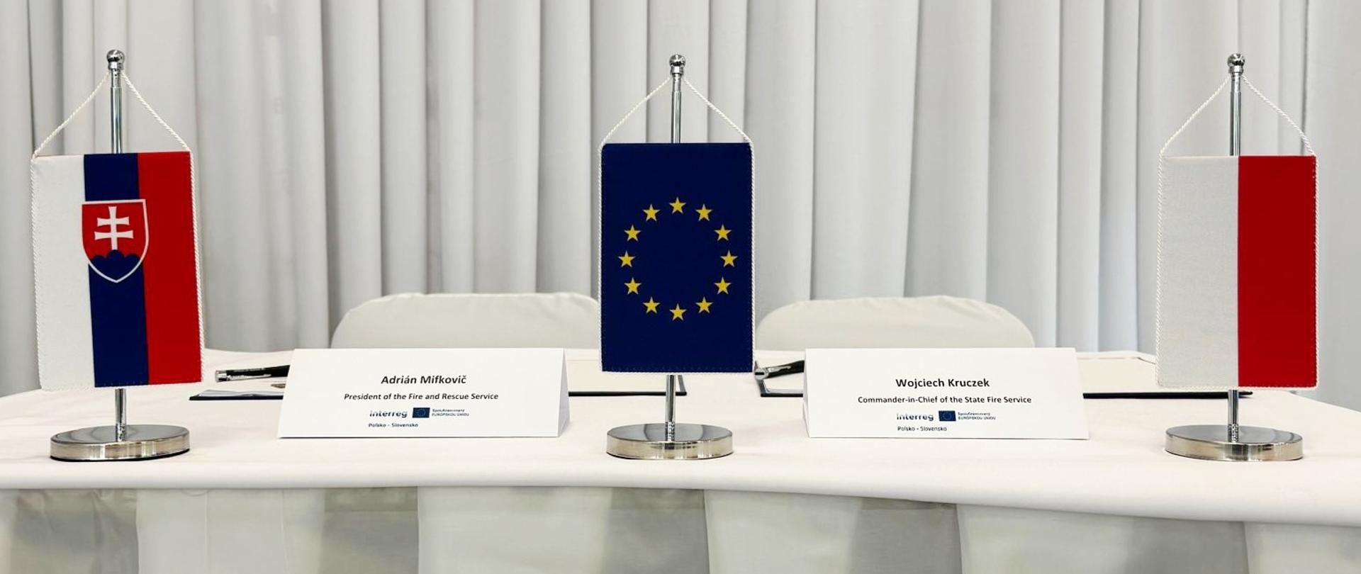 Stół konferencyjny przygotowany do podpisania dokumentu – ustawione trzy miniaturowe flagi: Słowacji, Unii Europejskiej i Polski oraz tabliczki z nazwiskami sygnatariuszy.