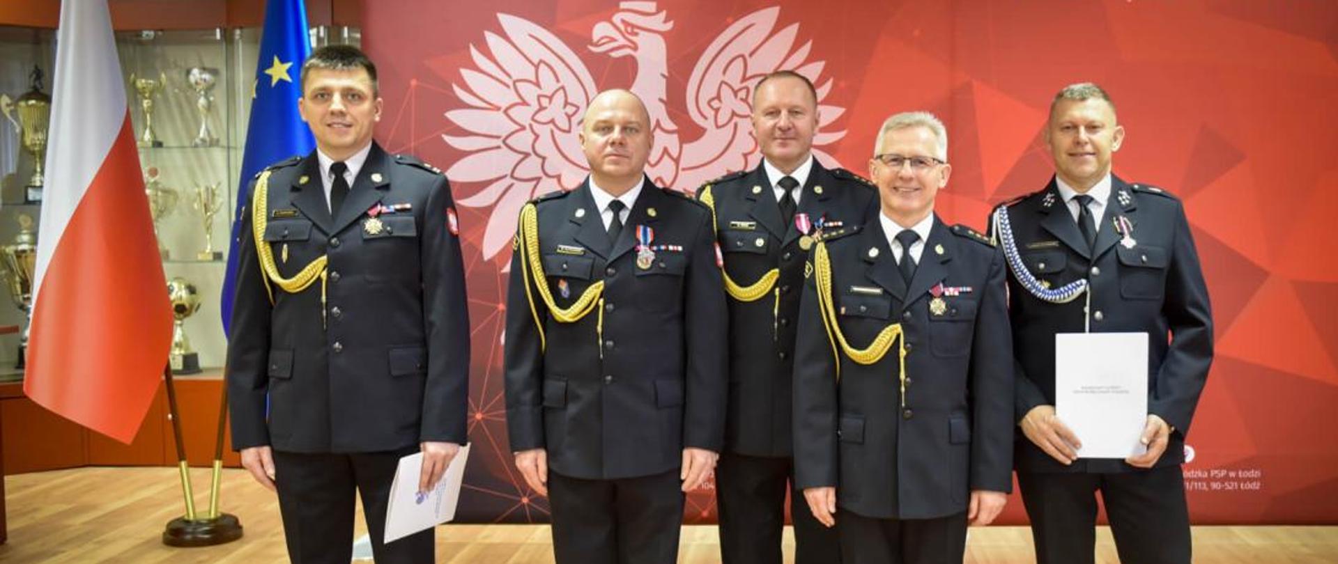 Funkcjonariusze Komendy Powiatowej Państwowej Straży Pożarnej w Brzezinach i druh Sławomir Młotkowski ubrani na galowo, na sali uroczystości pozują po wręczeniu odznaczeń i awansów na wyższe stopnie służbowe, które zostały nadane podczas uroczystości z okazji Narodowego Święta Niepodległości