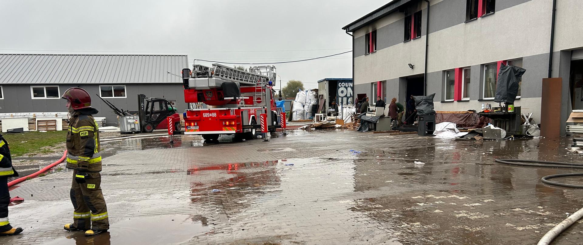 Funkcjonariusze Straży Pożarnej stoją na otwartej przestrzeni. W tle stoi wóz strażacki. Po prawej stronie widać jednopiętrową halę, która uległa spaleniu.