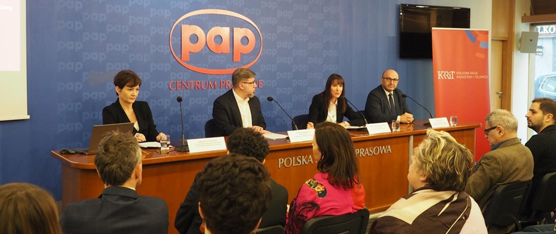 Prelegenci siedzący za stołem. W tle granatowa ściana z czerwonym napisem PAP.