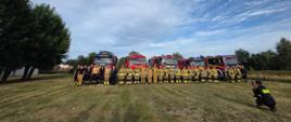 Grupa kilkudziesięciu strażaków w żółtych mundurach z elementami odblaskowymi pozuje na tle samochodów strażackich ustawionych na trawiastym terenie. Wokół terenu rosną zielone drzewa. Nad strażakami i pojazdami widoczne jest błękitne niebo z rozciągniętymi białymi chmurami. Przed grupą pozujących znajduje się strażak, który w półprzysiadzie patrzy w stronę strażaków trzymając w rękach czarny nieduży przedmiot podobny do smartfona. 