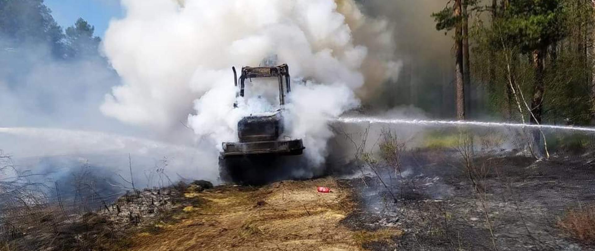 Na środku zdjęcia widać palącą się maszynę nad którą unoszą się kłęby białego dymu. Przed maszyną znajduje się droga leśna, na której leży zużyta gaśnica. Po obu stronach drogi widać pogorzelisko po palącym się poszyciu lasu. Z prawym górnym rogu zdjęcia widać rosnące drzewa sosnowe w górnej części zdjęcia widoczne są linie wysokiego napięcia, które przebiegają wzdłuż drogi leśnej. W lewej części zdjęcia widoczne są korony drzew lasu sosnowego.