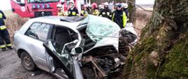 Zdjęcie przedstawia samochód marki Audi po uderzeniu w drzewo oraz strażaków i policjanta na tle samochodu pozarniczego