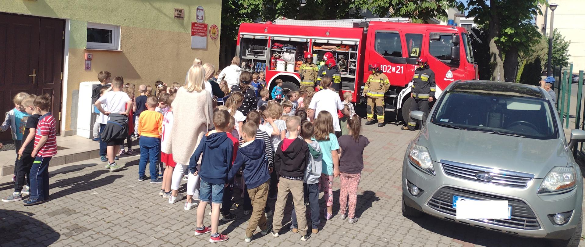 Na zdjęciu widać dzieci wraz z wychowawczyniami oraz strażaków ubranych w ubrania bojowe stojących na tle samochodu gaśniczego.