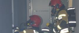 Ćwiczenia taktyczne jednostek ochrony przeciwpożarowej. Teren jednej z firm w Rawiczu. Dwóch strażaków znajduje się na korytarzu na drugiej kondygnacji budynku. Jeden z nich trzyma w rękach wąż z przykręconą prądownicą. Drugi klęczy przed drzwiami do pomieszczenia.
