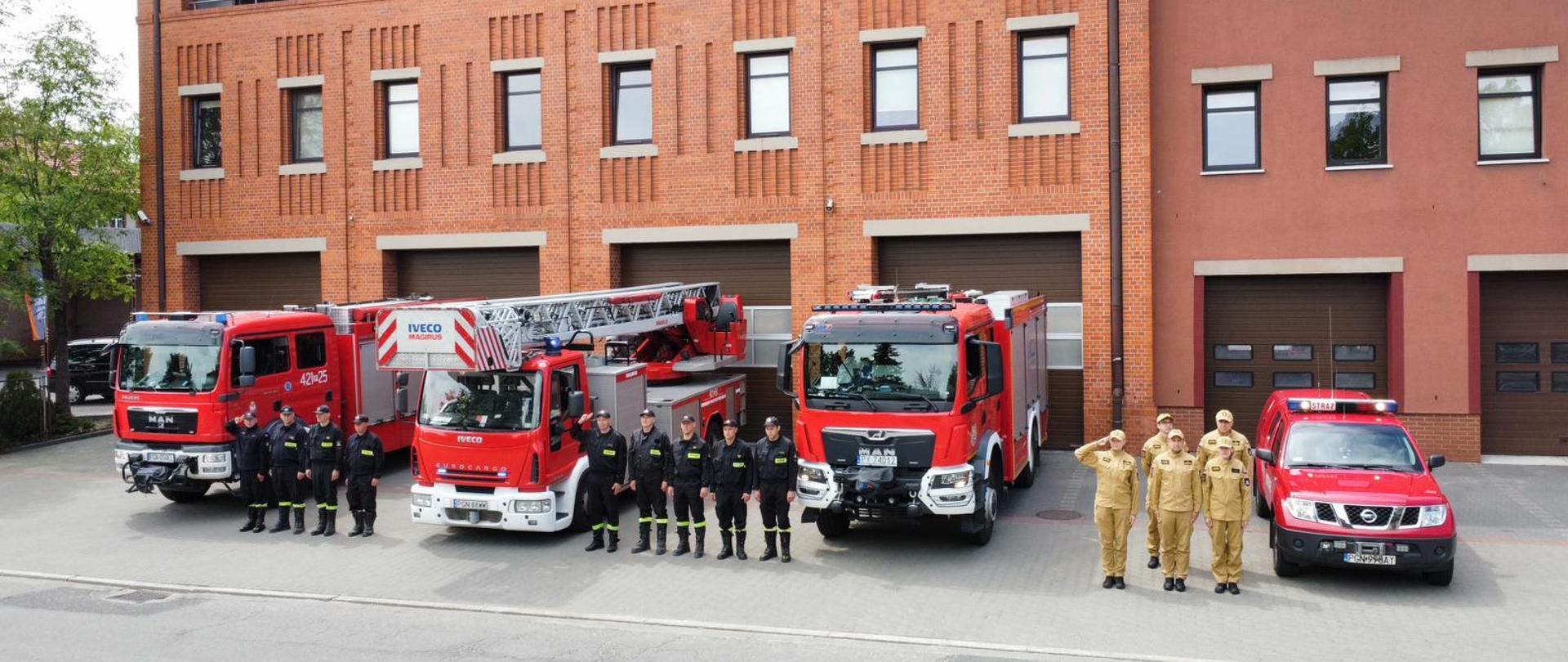 Strażacy stoją przed budynkiem z czerwonej cegły obok czerwonych pojazdów strażackich 
