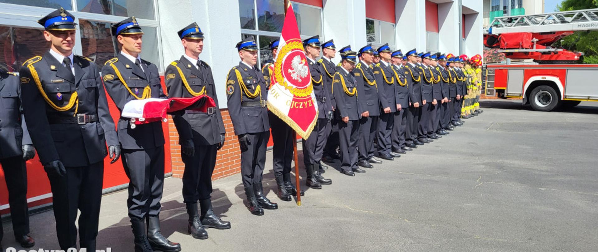 w dlugim szeregu stoją strażacy w ciemnych mundurach ze sznurem i w rogatywkach, na początku szeregu stoi poczet flagowyt oraz sztandarowy