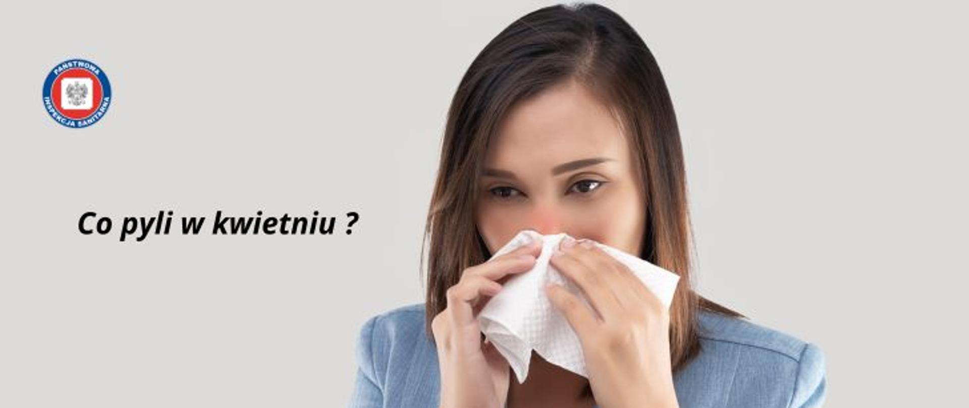 Na szarym tle kobieta w długich ciemnych włosach wycierająca nos białą chusteczką jednorazową. Po lewej stronie napis Co pyli w kwietniu? W lewym górnym rogu okrągłe logo Państwowej Inspekcji Sanitarnej przedstawiające czarnego orła na białym kwadratowym tle, które otoczone jest czerwonym wypełnieniem. Zewnętrzną część logo otacza granatowy okrąg z napisem Państwowa Inspekcja Sanitarna w kolorze białym.