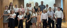 Na zdjęciu widać dużą grupę młodych ludzi, którzy są ubrani w odświętne stroje, po występie muzycznym. Stoją na podwyższeniu, uśmiechnięci.
Na pierwszym planie, na dole, stoi pięcioro dzieci, każde z nich trzymające flet poprzeczny.
W drugim rzędzie, po lewej stronie, stoi dziewczynka w jasnej sukience, a po jej prawej stronie stoi dziewczynka z fletem poprzecznym.
W głębi, po prawej stronie, stoi grupa chłopców z trąbkami
W tle, za dziećmi, stoi dorosła kobieta i jeden mężczyzna, nauczyciele. Na ścianie za nimi wisi obraz, który przypomina okno. Po lewej stronie widać drewniane panele na ścianie.
W lewym dolnym rogu zdjęcia widnieje data: "05 cze 2025".