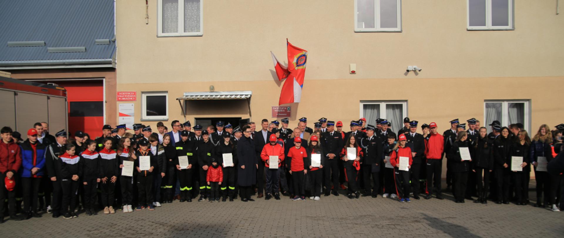 Zaproszeni goście pozujący do pamiątkowego zdjęcia. Zdjęcie wykonano w słoneczny dzień na placu przed remizą Ochotniczej Straży Pożarnej w Zbrzeźnicy.