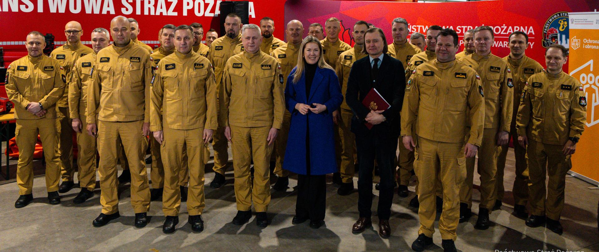Zdjęcie grupowe strażaków PSP oraz zaproszonych gości wykonane w garażu jednostki. W tle samochód pożarniczy i ścianka promocyjna PSP.