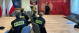 Strażacy ubrani w czarne ubrania koszarowe siedzą w ławkach przodem skierowani do stołu prezydialnego w trakcie szkolenia z zakresu współpracy z mediami. Na przodzie stoją strażacy prowadzący szkolenie, widać flagi Polski godło Polski oraz ściankę KM PSP Tarnów