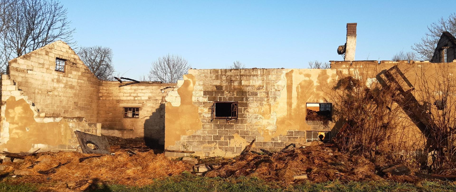 Na zdjęciu widać spalony budynek gospodarczy bez dachu. Przed budynkiem leżą nadpalone rzeczy wyciągnięte z wnętrza budynku. Budynek nie ma drzwi oraz jedna ze ścian jest częściowo zawalona. Ściany są zbudowane z białego pustaka. W tle widać koronę drzew bez liści. Jest widno.